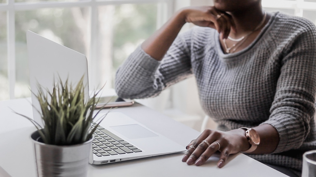 A black business woman sitting in front of window wearing a gray sweater hosting a virtual event. This is a way to still do business during social distancing  as a result of coronavirus aka covid-19. 